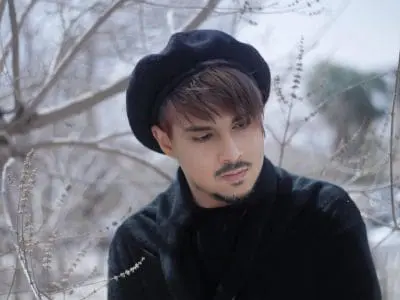 Portrait of a man with brown hair wearing a black beret and dark coat in a snowy, bare-branch setting.