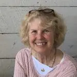 Smiling older woman with short blonde hair, pink knit sweater, glasses on her head, against a light wooden wall background.