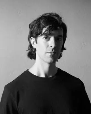 Black-and-white portrait of a young man with tousled dark hair, wearing a dark T-shirt, standing against a plain textured wall.