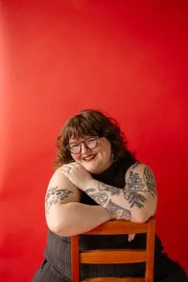 Smiling woman with tattoos leaning on a wooden chair against a solid red background.