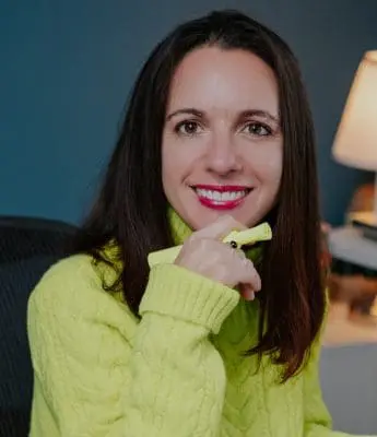 Portrait of a smiling woman with long dark hair wearing a bright lime sweater, holding a yellow highlighter near her chin in a teal-walled room.
