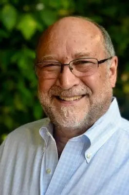 Smiling older man with glasses and a light blue button-down shirt, outdoors with green foliage in the background.