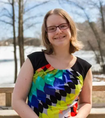 Smiling woman wearing a colorful patterned dress sits on a wooden bench outdoors with bare trees in the background.