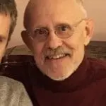 Close-up of a smiling elderly man with glasses, balding on top and white beard, wearing a burgundy turtleneck indoors.